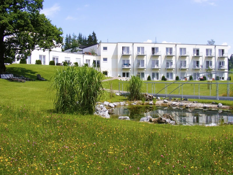 Au&szlig;enansicht der Oberberg Fachklinik Scheidegg im Allg&auml;u: helles Klinikgeb&auml;ude vor bewaldeten H&uuml;geln und blauem Himmel